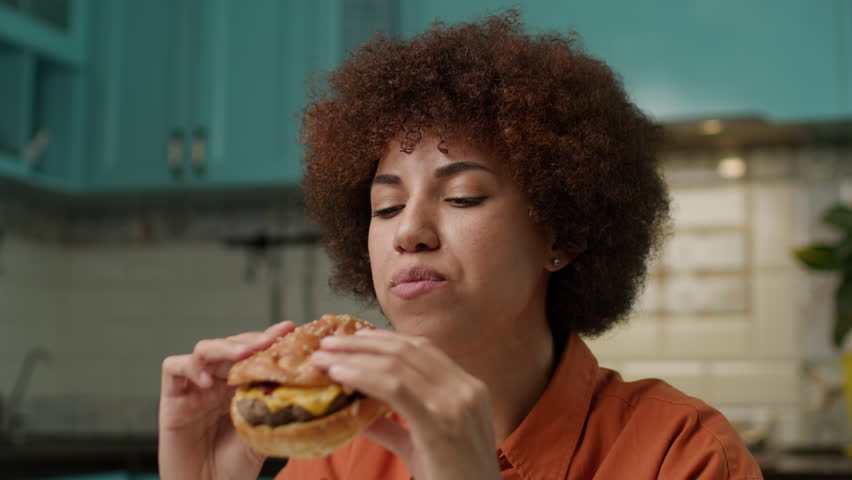 Black Woman Eating Yummy Burger Close Up. 20s female enjoy cheeseburger looking at camera. Royalty-Free Stock Footage #1105764587