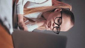 Vertical video of dreamy preteen girl dressed in white blouse and black glasses sitting still in office chair and slowly typing on keyboard. Blue-eyed child holding hand under chin and thinking.