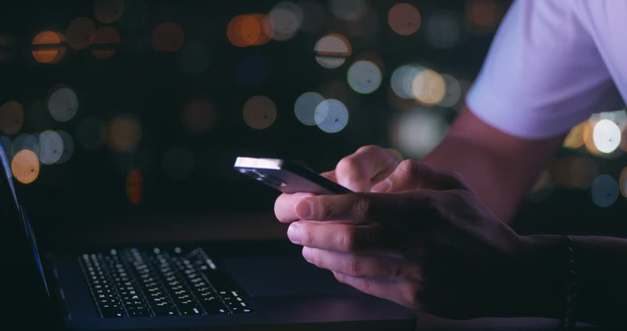 Close-up of male hands hold mobile phone, texting message while working on a laptop computer on background of a night city. Man freelancer using a mobile phone while working at computer in the evening Royalty-Free Stock Footage #1105769277