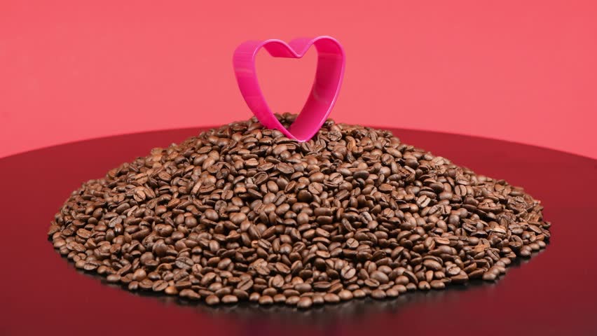 A red heart on a big pile of roasted coffee beans. Composition in red and black tones. The concept of coffee adoration. Side view. 4K UHD video footage 3840X2160.
