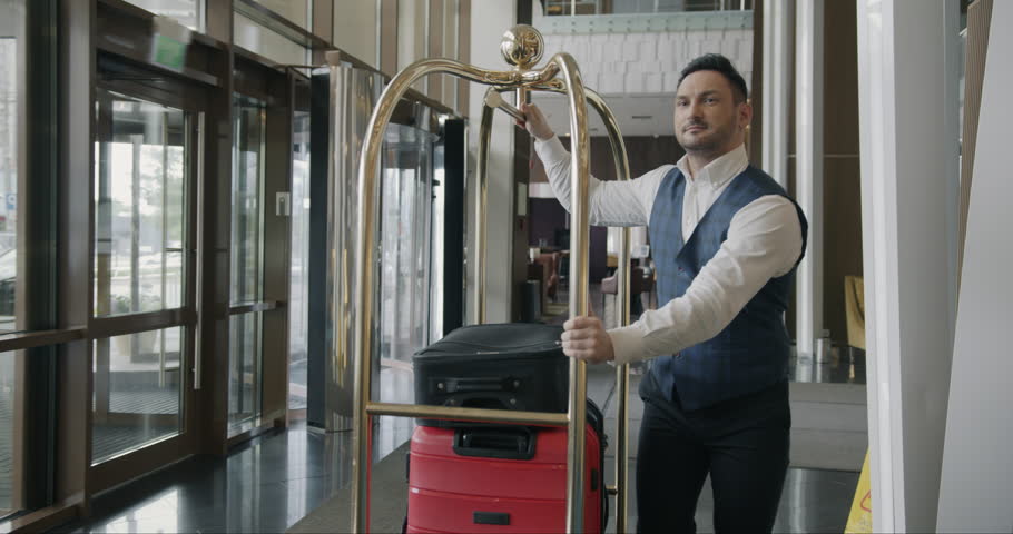 Dolly shot of helpful bellman pushing trolley with luggage along hotel corridor working alone wearing uniform. People and occupation concept. Royalty-Free Stock Footage #1105770109