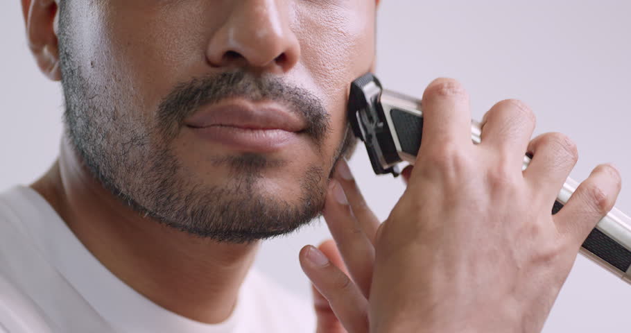 A man is using electric shaver to trim mustache and beard while standing in front of mirror in bathroom at home.