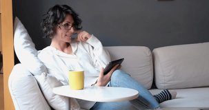 Smiling woman motivating at video call.Young brunette woman with curly hair and casual clothing sitting on the sofa and looking, chatting, reading on her notepad. Horizontal indoor video in 4k resolut