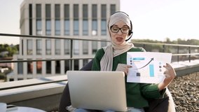 Young arabian female in casual clothes and hijab giving online presentation using headset and computer outside office. Remote manager brainstorming about statistics shown by infographics on terrace. - Powered by Shutterstock - Get 15% off with code: PIKWIZARD15