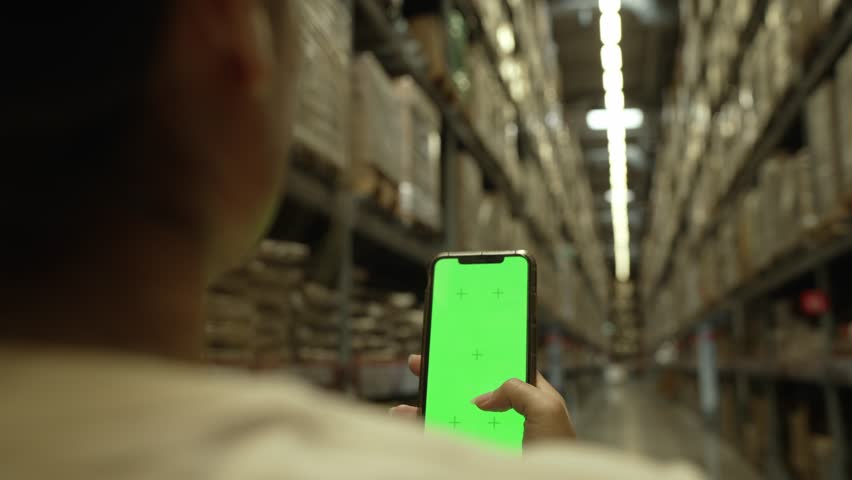 Woman's hand touching a green screen, scrolling, browsing for products on a smartphone in a large warehouse. Royalty-Free Stock Footage #1105781649