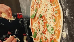 Hands spreading grated cheese on pizza. Vertical video. Cooking pizza at home. Woman preparing italian homemade pizza, adding cheese on fresh dough. Process of making delicious healthy food