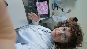 Vertical shot of female Caucasian scientist making vertical video showing research results in laptop in laboratory at daytime