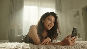 Attractive brunette communicating through video call lying on bed on stomach in bedroom lit by sun rays. Young woman is pleasantly surprised, happy. High quality 4k footage