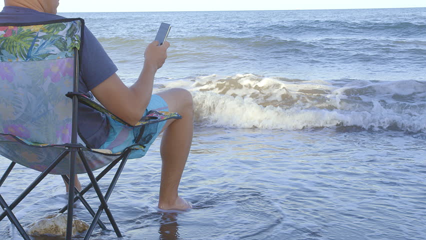 Man resting in a portable chair on the seashore, waves touching his feet, man sitting with cell phone in hand Royalty-Free Stock Footage #1105790437