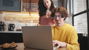 Attractive man using laptop while his beloved woman standing behind with glass of water. People spending time at modern kitchen. Choosing future purchase online. Concept of Internet. - Powered by Shutterstock - Get 15% off with code: PIKWIZARD15