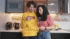 Couple standing in kitchen while watching funny videos on smartphone. Young people spending time together. Pretty woman holding cup of drink and smiling together with her husband. Enjoying rest.