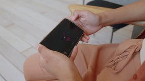 Close-up arc shot of hands of anonymous young woman sitting in chair holding smartphone with media player app on screen, in minimalist beige interior