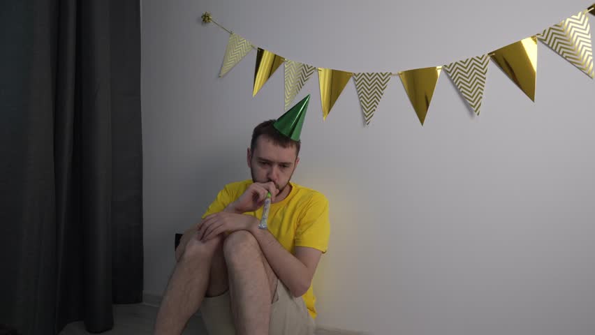 Sad upset Caucasian young man alone in with a party cap in celebration atmosphere. Guy angry sitting on the floor visually sad about something slow motion Royalty-Free Stock Footage #1105792491