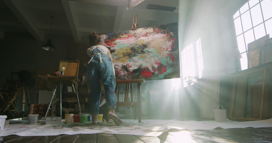 Millennial girl artist paints picture in loft studio using her hands to apply paint to canvas. Abstraction cinematic. Talented female artist energetically creates masterpiece of painting. light rays Royalty-Free Stock Footage #1105797941