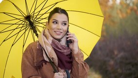 Caucasian young woman on a phone call holding a yellow umbrella smiling in autumn.