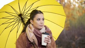 Caucasian young woman with coffee and yellow umbrella looking at camera and smiling in autumn.