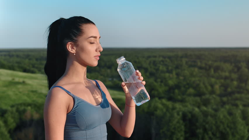 Sport brunette woman drinking fresh pure mineral water from bottle outdoor on mountain over forest sky panorama. Athletic female enjoy aqua beverage with thirst healthy lifestyle at nature landscape Royalty-Free Stock Footage #1105806345