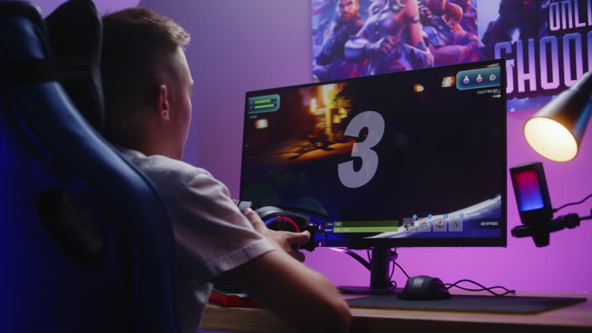 Schoolboy puts on headphones and starts play third person shooter. Gamer enjoys online video game on computer at home. Desk illuminated by RGB LED strip light. Concept of gaming at home. Dolly shot.