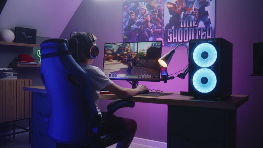 Young gamer plays third person shooter on personal computer. Schoolboy in headphones enjoys online video game at home. Desk illuminated by RGB LED strip light. Concept of gaming at home. Zoom in.