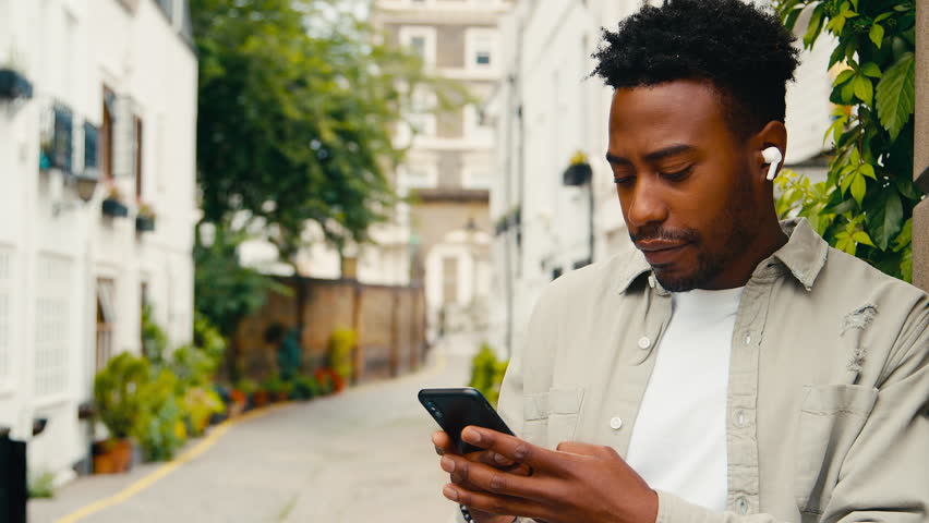 Young man travelling through city listening to music on wireless earbuds on mobile phone - shot in slow motion