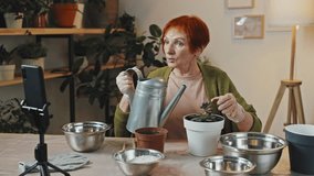 Panning right medium shot of senior woman in green cardigan sitting at table making video and watering plant