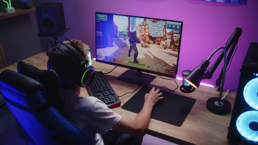 Young gamer plays third person shooter on personal computer. Schoolboy in headphones enjoys online video game at home. Desk illuminated by RGB LED strip light. Concept of gaming at home. Dolly shot.