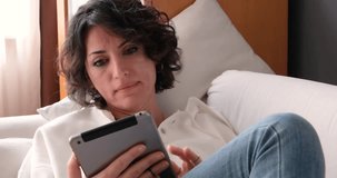 Young brunette woman with curly hair and casual clothing is sitting on the sofa and looking, chatting, reading, watching on her notepad. Horizontal indoor video in 4K resolution of female person