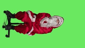 Vertical video Front view of pensive santa in costume sits on chair, thinking about christmas eve gift ideas over full body greenscreen. Young man acting like saint nick character with white beard
