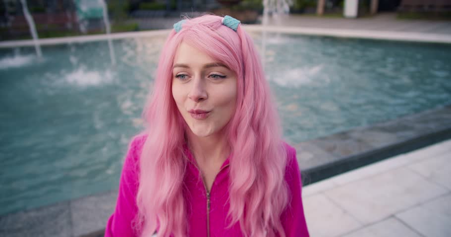 A girl in pink is sitting near the fountain, having fun and inflating a bubble gum balloon. Cheerful bright young girl Royalty-Free Stock Footage #1105820435