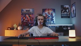 Professional teenage gamer in headphones speaks into microphone, plays online video game on computer at night in stylish neon room. ESports tournament. Gaming at home. View from PC screen perspective.