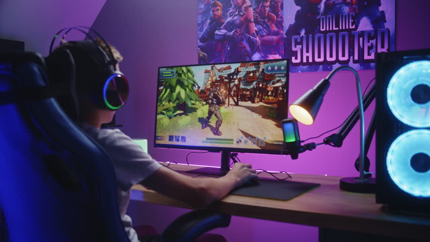 Young gamer plays third person shooter on personal computer. Schoolboy in headphones enjoys online video game at home. Desk illuminated by RGB LED strip light. Concept of gaming at home. Back view.