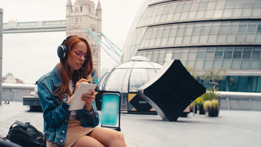 Female vlogger or social influencer wearing headphones travelling through city writing in notebook - shot in slow motion