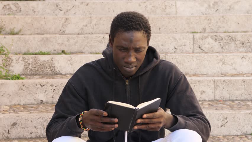 Portrait of young african man sitting on the street concentrating on reading the notes in the diary | Shutterstock HD Video #1105826893