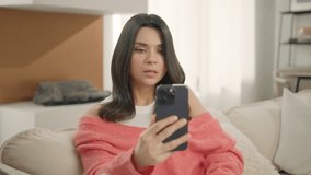 Happy young brunette woman using smart phone camera and app with beautifying fashion funny face mask. Beautiful Social Media blogger looking at cell screen front camera, taking selfie sit on sofa home