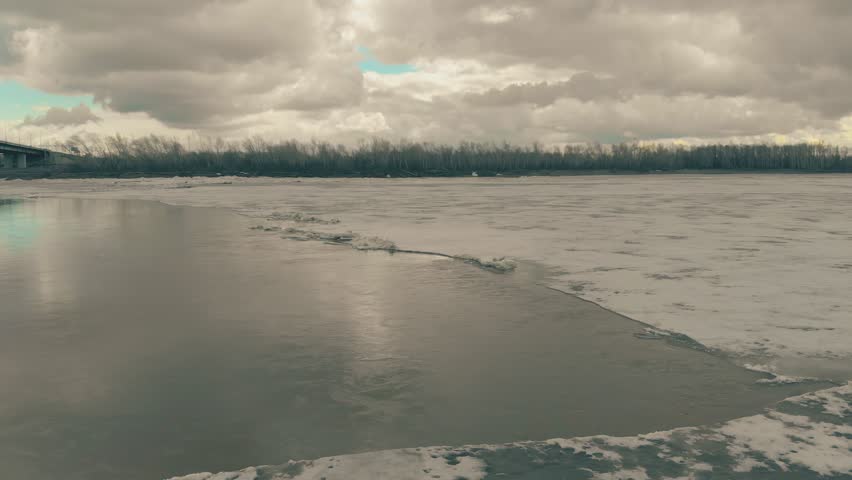 pictorial frozen river with patch of open water in ice against bank with dense forest bridge under cloudy sky upper view
