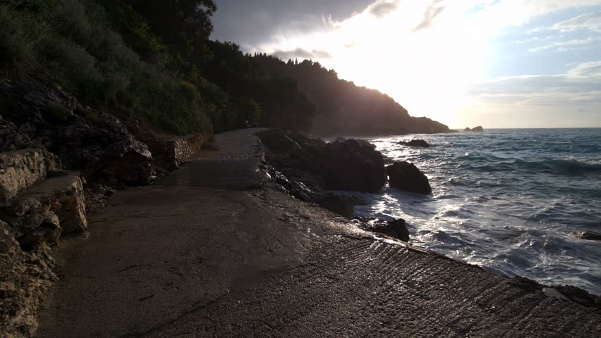 A walking tour of a foot path by the sea with strong waves and the sun setting behind the hill.