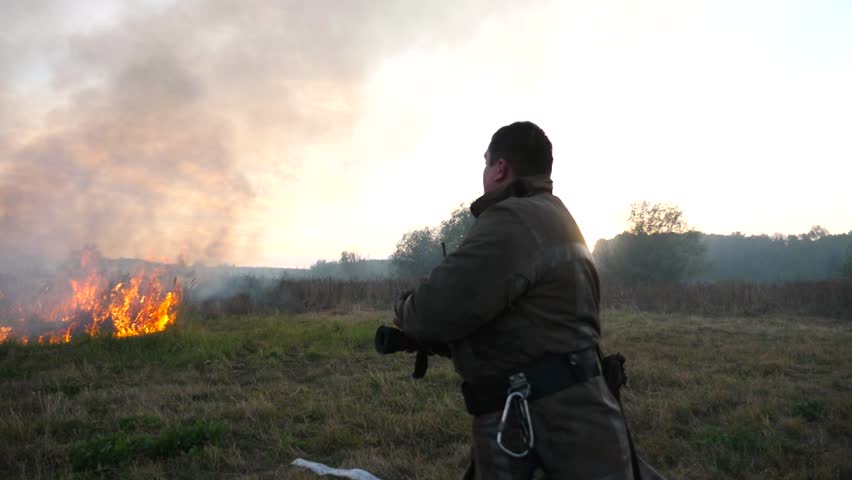 Male fireguard in equipment walking with fire hose on dry grass at countryside. Young fireman in uniform going to extinguish fire at meadow. Concept of environmental issues and global warming. Slow mo