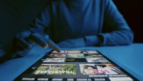 Close up of unrecognizable woman sitting at table with tablet lying down on it choosing film to watch on streaming service at night time