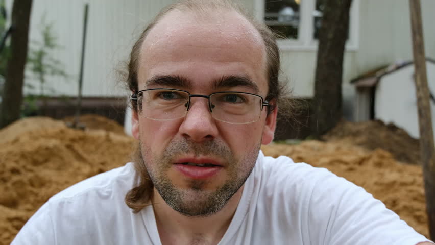 Close up of young adult man with facial hair and ponytail while wearing glasses and against pile of sand.