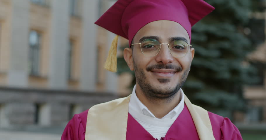 Slow motion portrait of cheerful Middle Eastern man student wearing gown and mortarboard standing outdoors in front of university smiling looking at camera Royalty-Free Stock Footage #1105870871