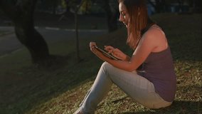 A beautiful woman sits in the park, sending some messages to her friends and family using her tablet.