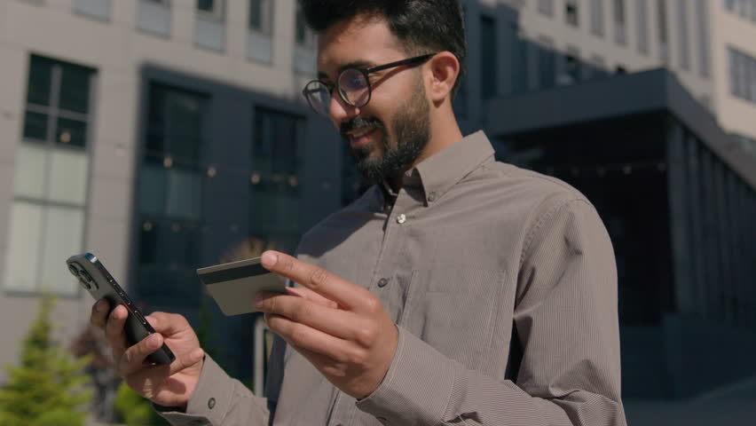 Happy Indian Arabian man hold mobile phone and credit bank card in city outdoors pay money online banking win good offer excited businessman celebrate business victory internet shopping achievement Royalty-Free Stock Footage #1105878347