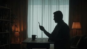 A man speaks on a video call using a telephone while standing at a table in the living room. Man waving his hand in greeting. Side view of the dark silhouette of a man.