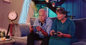Happy Indian two excited smiling old pensioner couple playing video game hold gamepad in hand sitting on sofa having funny competition moment spend weekend time enjoy together late night indoor home
