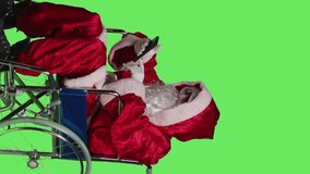 Vertical video Side view of man on phone call and playing videogames while he sits in wheelchair with seasonal festive costume, deal with physical disability. Santa claus character enjoys gaming