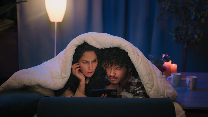 Couple Indian man and Arabian Hispanic woman night dating in bed under blanket family boyfriend girlfriend shocked watching horror film movie scared afraid frightened video with mobile phone hiding Royalty-Free Stock Footage #1105905219