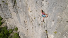 AERIAL: Young climber approaching the top of climbing route in limestone wall. Sporty woman clips belay rope in quickdraw while she climbs up the rocky wall. Exceptional location for sport climbing.
