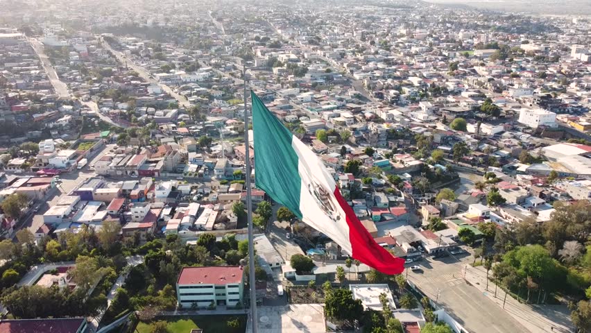 Beautiful aerial shot of Mexican flag waving over the border city of Tijuana. The camera pulls back gently and tilts up, revealing the horizon at around sunset.