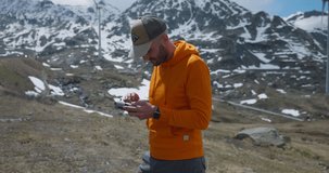 Caucasian bearded man standing while holding and setting parameters on drone remote controller with snow-capped mountains and wind turbine on background