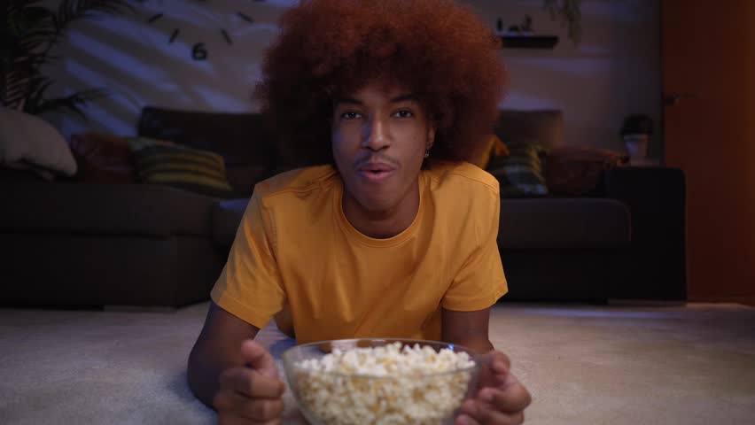 Point of view of an African American male teenager relaxed watching tv at home and eating popcorns while lying on the living room carpet.